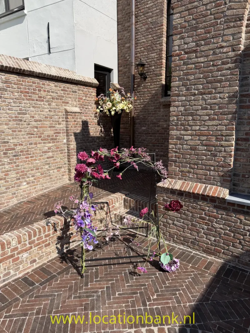 Bakstenen binnenplaats met een metalen boog vol bloemen en bloeiende planten in potten.