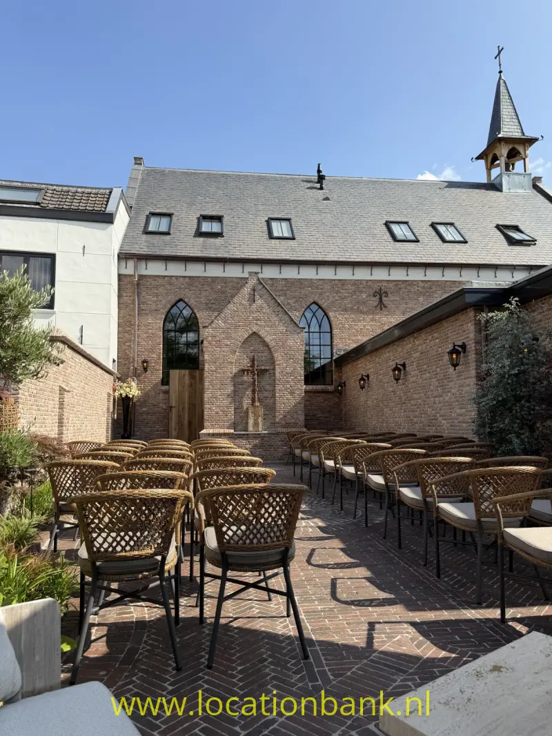 Bakstenen kerkgebouw met een leistenen dak, spits torentje en terras met gevlochten stoelen en tafels.