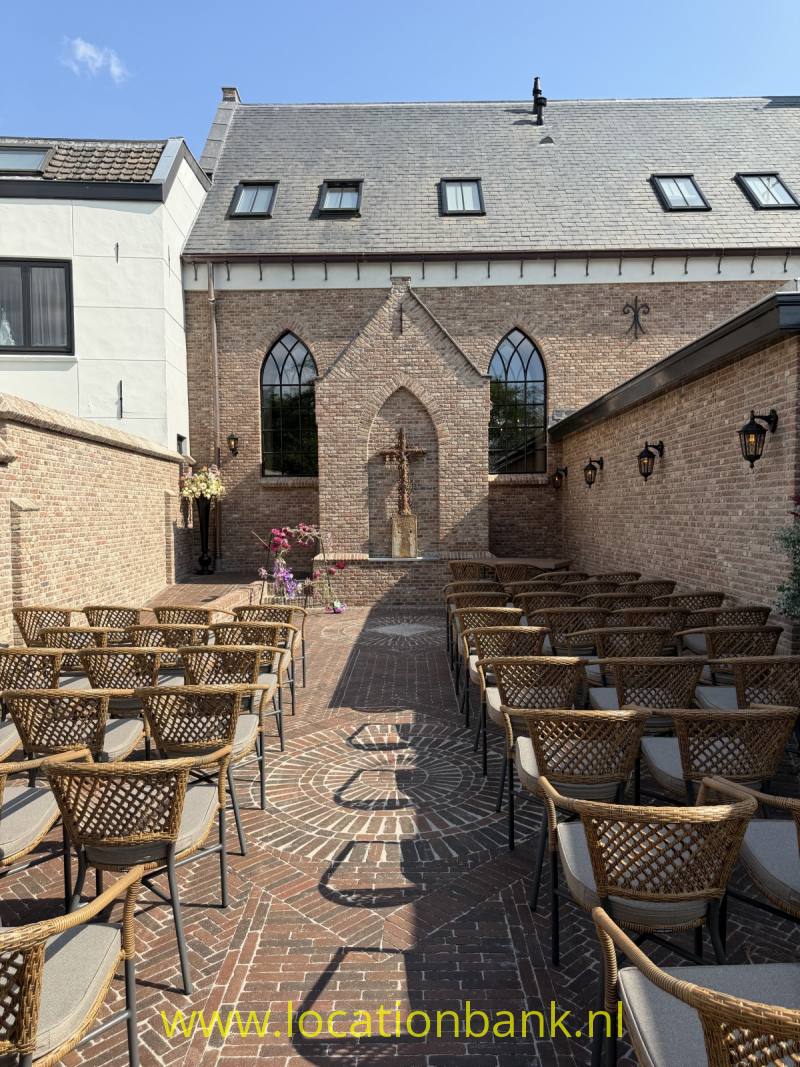 Bakstenen binnenplaats met rijen gevlochten stoelen en tafels, een puntgevel en hoge spitsboogvensters.