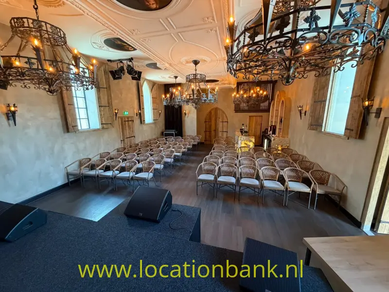 Rijk versierde zaal met rijen stoelen, kroonluchters, houten vloer en een verhoogd podium met luidsprekers.