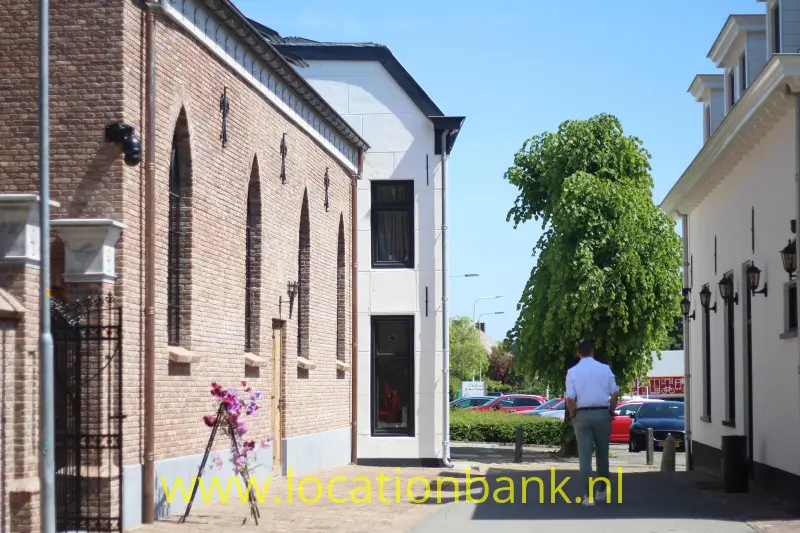 Bakstenen gebouw en witte panden staan langs een smalle straat met een boom, voetganger en geparkeerde auto’s.
