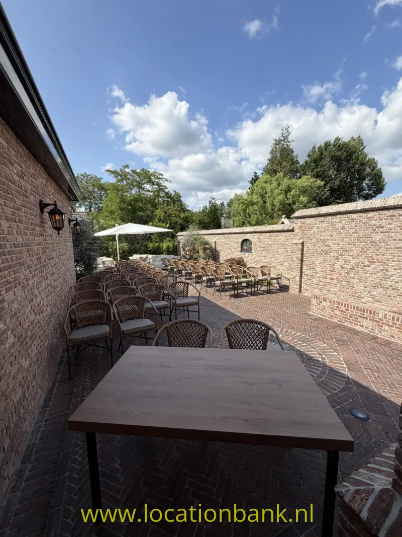 Bakstenen binnenplaats met tafels, rieten stoelen en parasols onder een blauwe hemel.