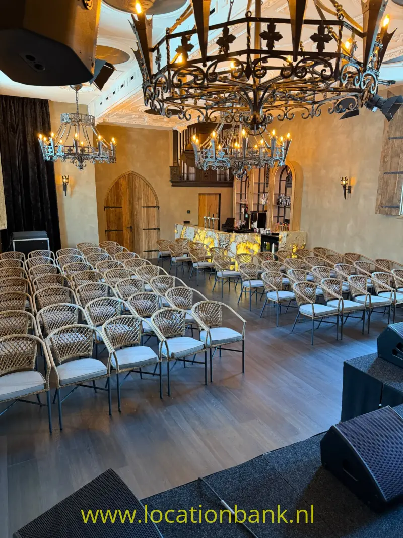 Rijen gevlochten stoelen staan in een zaal met meerdere kroonluchters, wandlampen en een houten boogdeur.