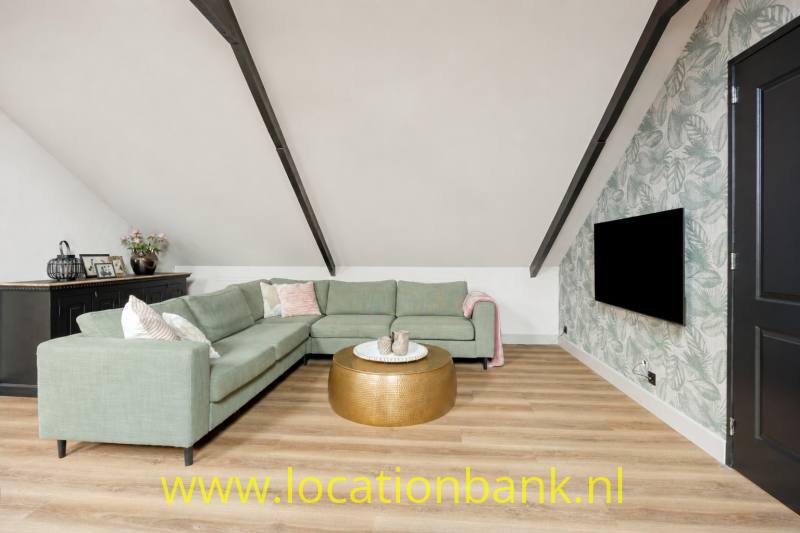 Een groene L-vormige hoekbank staat onder een schuin plafond, naast een zwart televisiescherm en botanisch behang.