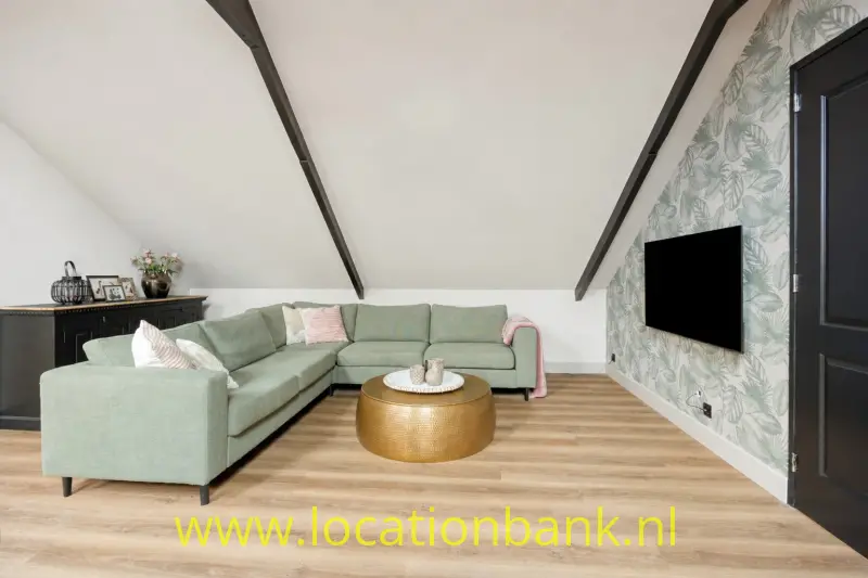 Een groene L-vormige hoekbank staat onder een schuin plafond, naast een zwart televisiescherm en botanisch behang.