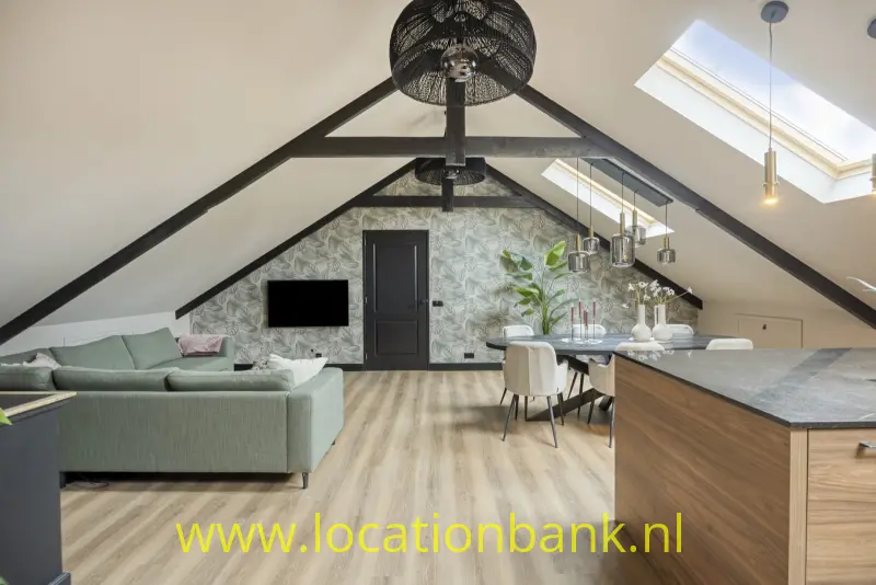 Open leefruimte onder een schuin dak met donkere balken, zithoek en keuken met kookeiland.