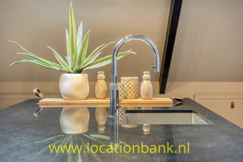 Een hoge chromen keukenkraan staat boven een spoelbak, met een plantenpot en houten serveerplank erachter op een donker werkblad.