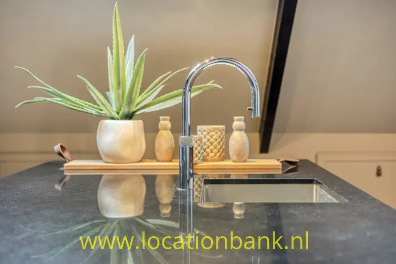 Een hoge chromen keukenkraan staat boven een spoelbak, met een plantenpot en houten serveerplank erachter op een donker werkblad.