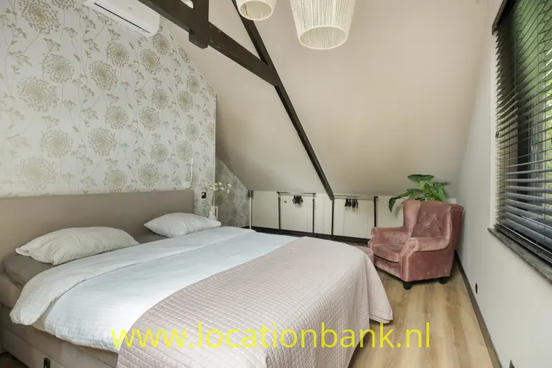 Slaapkamer met een opgemaakt tweepersoonsbed, bloemenbehang, donkere plafondbalken, een fauteuil en jaloezieën voor het raam.