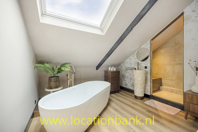 Vrijstaand wit bad onder een dakraam in een badkamer met een vloer in houtlook en een inloopdouche.