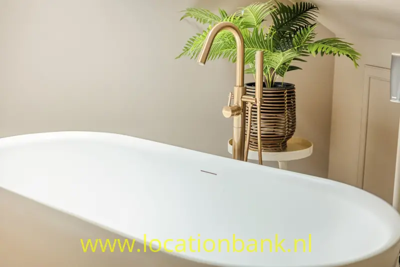 Vrijstaand wit bad met een gebogen goudkleurige kraan en hoge groene planten in een gevlochten mand.