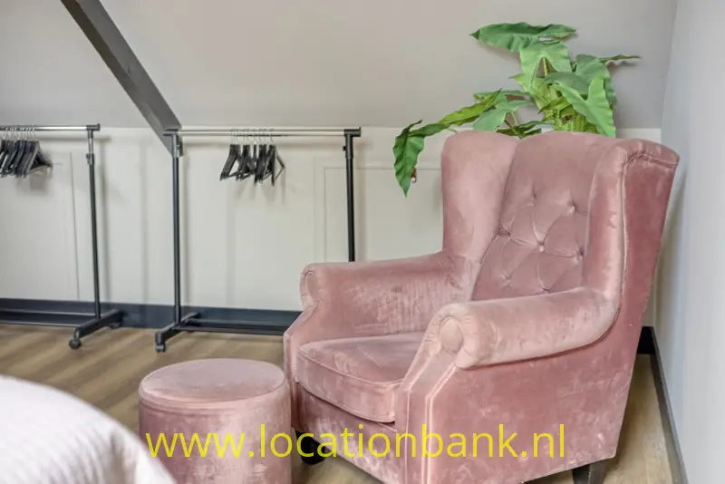 Een roze fauteuil met gecapitonneerde rugleuning staat naast een roze poef, met een groene plant erachter.