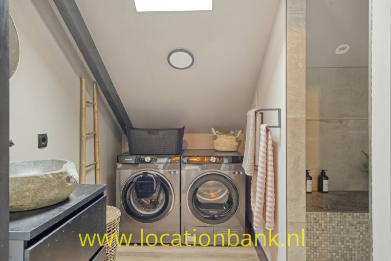 Een wasmachine en droger met voorlader staan onder een schuin plafond, naast een stenen wastafel en betegelde douche.
