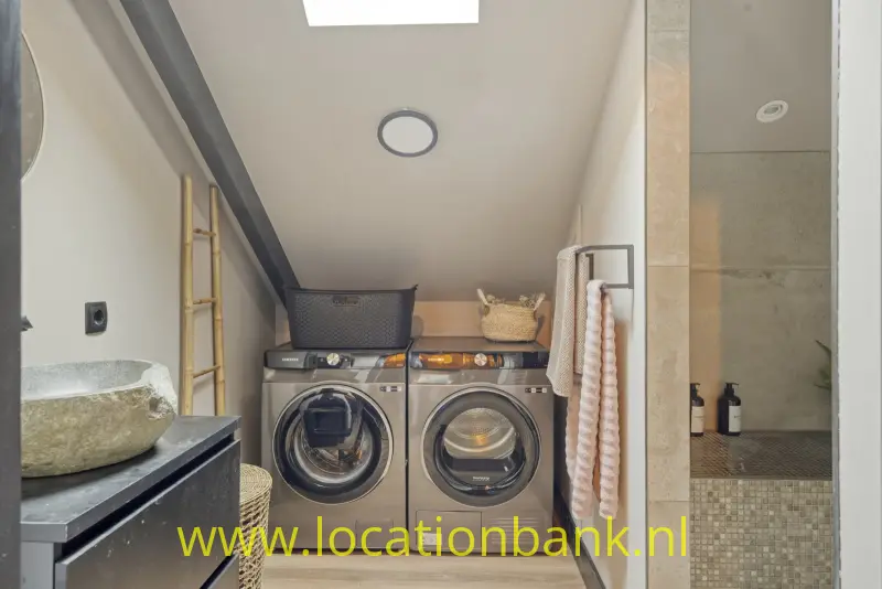 Een wasmachine en droger met voorlader staan onder een schuin plafond, naast een stenen wastafel en betegelde douche.