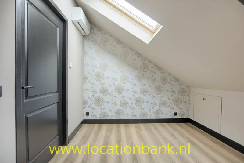 Kamer met een schuin plafond, dakraam, donkere deur, vloer in houtlook en behang met een bloemmotief.