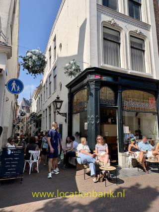 Verzierte Ladenfront mit hohen dekorierten Fenstern, Sitzplätzen im Freien und Fußgängern in einer schmalen, sonnigen Straße.