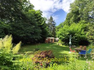 Ein grüner Garten mit hohen Bäumen, blühenden Pflanzen, einem hölzernen Gartenhaus und einer Sitzecke mit Sonnenschirm.