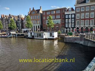 Ein Hausboot liegt in einem Kanal, dahinter stehen schmale hohe amsterdam Häuser und eine steinerne Kaimauer.