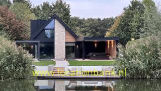 Modernes schwarzes Haus mit hoher Steinfassade, einer Terrasse am Wasser und einem Teich im Vordergrund.