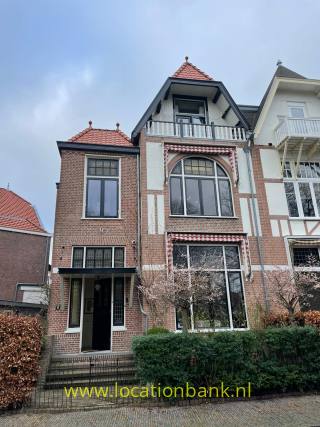 Backsteinhaus mit roten Ziegeldächern, weißen Balkonen und großen Rundbogenfenstern hinter einer Hecke.