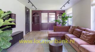 Wohnzimmer mit einem rosafarbenen Ecksofa, runden Holzcouchtischen, großen Pflanzen und dunklen Holzschränken.