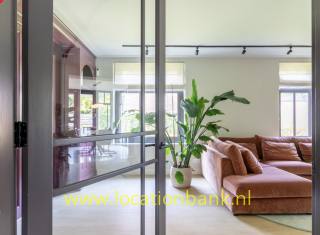 Wohnzimmer mit einer Glaswand, einem braunen Ecksofa, einer großen Zimmerpflanze und Schienenbeleuchtung.
