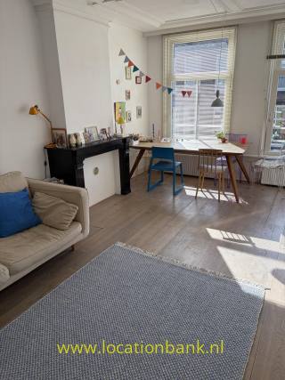 Wohnzimmer mit einem großen Fenster mit Jalousien, einem Schreibtisch, zwei Stühlen, einem Sofa und einer schwarzen Kaminverkleidung.