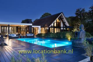 Reetgedecktes Haus mit verglaster Giebelfassade, beleuchtetem Swimmingpool, Terrasse und Skulptur in der Dämmerung.