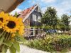A house with black wooden siding, red roof tiles and sunflowers stands beside a flowering garden.
