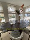Oval dark dining table with a vase of branches, glasses and chairs in front of tall windows.