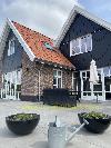 Dark façades with white frames, red roof tiles, a terrace and planters with a watering can in the foreground.