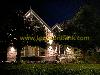 Illuminated brick house with gables, surrounded by trees and a low hedge beneath a dark night sky.