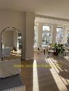 Living room with tall bay windows, a round mirror, a dining table and chairs on a wooden floor.