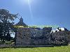 Großes Schloss mit einem Türmchen, umgeben von einer Rasenfläche und hohen Bäumen unter klarem Himmel.