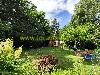 Ein grüner Garten mit hohen Bäumen, blühenden Pflanzen, einem hölzernen Gartenhaus und einer Sitzecke mit Sonnenschirm.