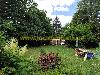 Grüner Garten mit einer Rasenfläche, Blumenbeeten, Gartenmöbeln und eines kleines Holzhauschen zwischen den Bäumen.