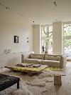 Stimmungsvolles, geräumiges Wohnzimmer mit einer sanften, natürlichen Ausstrahlung. Ein großzügiges helles Sofa, ein niedriger Couchtisch aus Holz, Wollteppiche und dezente Kunstobjekte verleihen dem Raum eine zeitgemäße, ruhige Atmosphäre. Große Fenster sorgen für viel Tageslicht und bieten einen Blick auf die Straße und das Grün im Außenbereich.