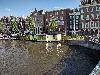 Ein Hausboot liegt in einem Kanal, dahinter stehen schmale hohe amsterdam Häuser und eine steinerne Kaimauer.