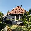 Schwarzes Holzhaus mit rotem Ziegeldach, zwei Fenstern und einer Bank im Garten.