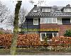 Haus mit dunkler Fassadenverkleidung, einer Backsteinfassade, Gauben, Dachfenstern, einer Hecke und einem Baum.