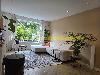 Wohnzimmer mit einem weißen Ecksofa, einem großen Fenster mit Vorhängen und grünen Pflanzen.