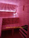Holzverkleideter Sauna mit mehreren Sitzbänken, horizontalen Wandbrettern und einem kleinen Fenster unter rosafarbener Beleuchtung.
