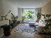 Helles Wohnzimmer mit großen Fenstern, einem Ecksofa, Zimmerpflanzen, einem Teppich und einem Fischgrätboden.