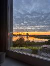 Ein von Schilf gesäumter See spiegelt den orangefarbenen Sonnenuntergang, gesehen durch ein Fenster.
