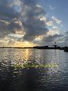 Eine Windmühle steht am Wasser, während die tief stehende Sonne durch dunkle Wolken scheint.