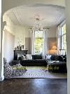 Wohnzimmer mit dunklen Sofas, hohen Fenstern, einem Kronleuchter und dekorativem Deckenstuck.