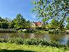 Ein Haus mit roten Dächern liegt hinter einem Teich, umgeben von Bäumen und Schilf.