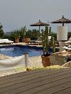 Swimmingpool mit Liegen, Sonnenschirmen, Kakteen, einer Außenleuchte und einer Holzterrasse im Vordergrund.