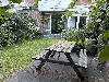 Ein hölzerner Picknicktisch steht auf einer gepflasterten Terrasse in einem grünen Innenhof neben einem Ziegelgebäude.