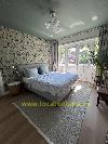 Schlafzimmer mit einem bezogenen Bett, botanischer Tapete, hohen Fenstern, Vorhängen und einer großen Zimmerpflanze.